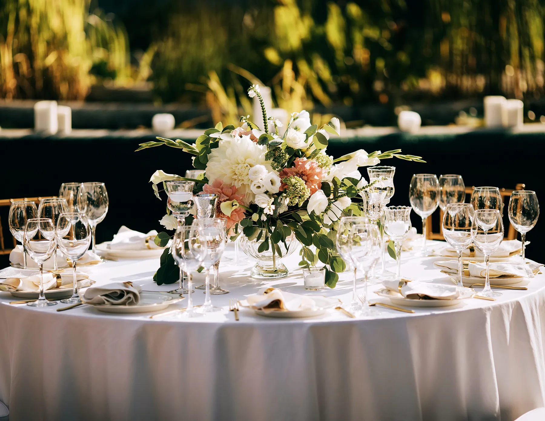 outdoor-wedding-reception-table-with-peach-and-white-floral-centerpiece-hmg-plus. An elegantly set outdoor wedding reception table with a white linen, crystal glassware, gold cutlery, and a lush centerpiece of peach and white blooms with eucalyptus greenery.