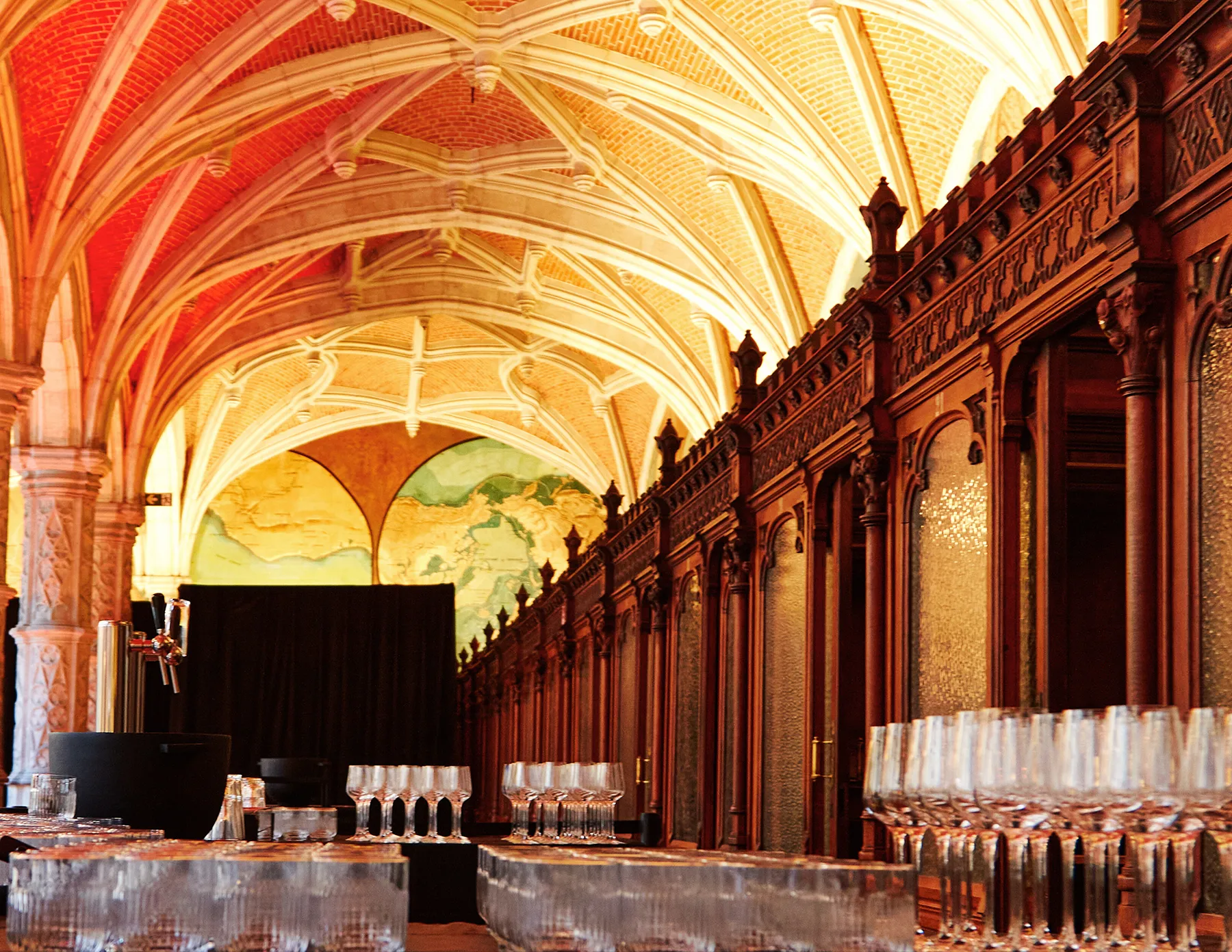 historic-gothic-vaulted-hall-set-up-as-event-bar-hmg-plus. Rows of crystal glassware and beer taps line a bar setup inside a magnificent Gothic-vaulted hall with ornate dark wood paneling and a large world map mural illuminated at the far end.