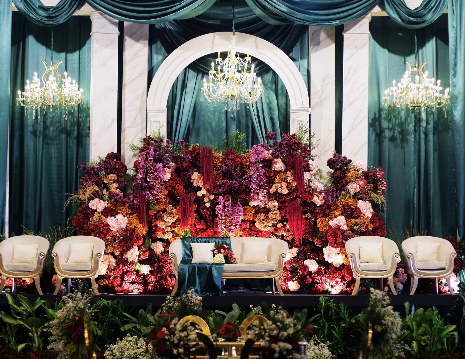 luxury-floral-wedding-stage-teal-burgundy-hmg-plus. An opulent wedding reception stage featuring an abundant floral wall backdrop of burgundy, deep purple, pink, and amber blooms, framed by a white marble arch and dramatic teal satin drapes, with cream upholstered chairs and crystal chandeliers.