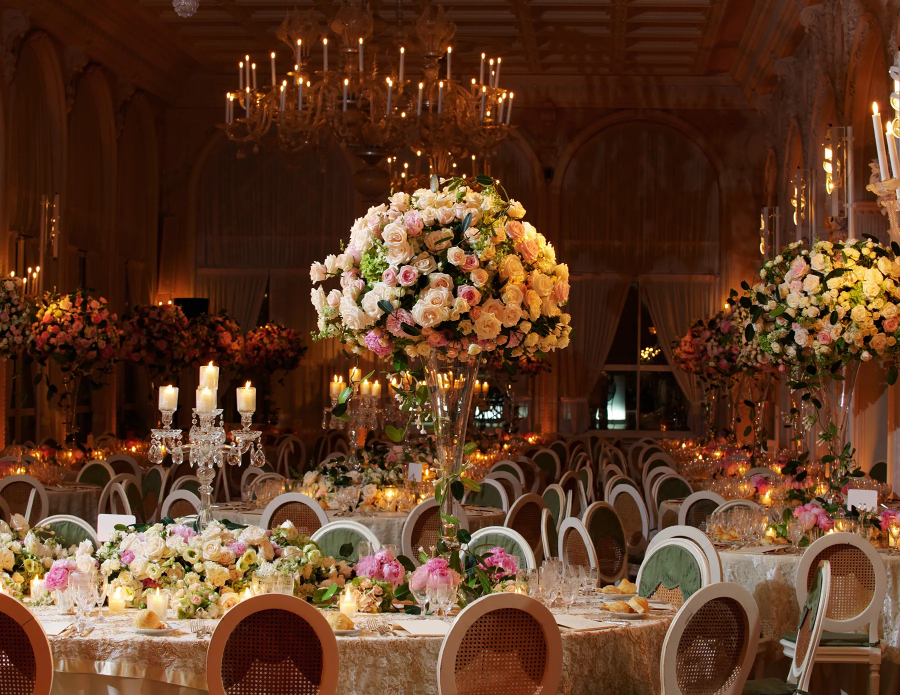 opulent-ballroom-wedding-reception-with-towering-floral-centerpieces-hmg-plus. A grand ballroom wedding reception featuring towering arrangements of peach, pink, and cream roses on candlelit tables, beneath an ornate crystal chandelier.