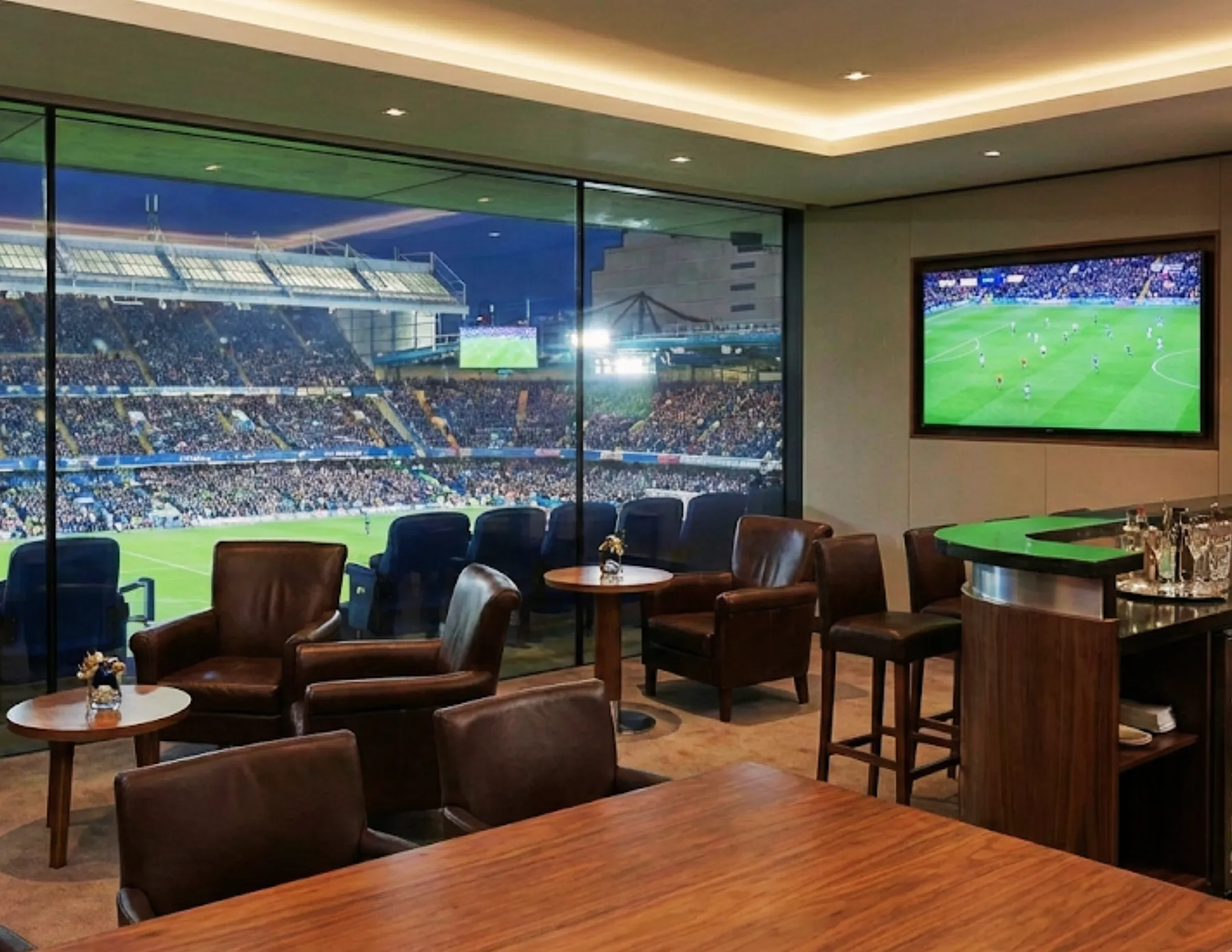 luxury-stadium-suite-with-pitch-view-hmg-plus. Luxury private stadium suite with lounge seating, bar area, and floor to ceiling glass overlooking a live sports event