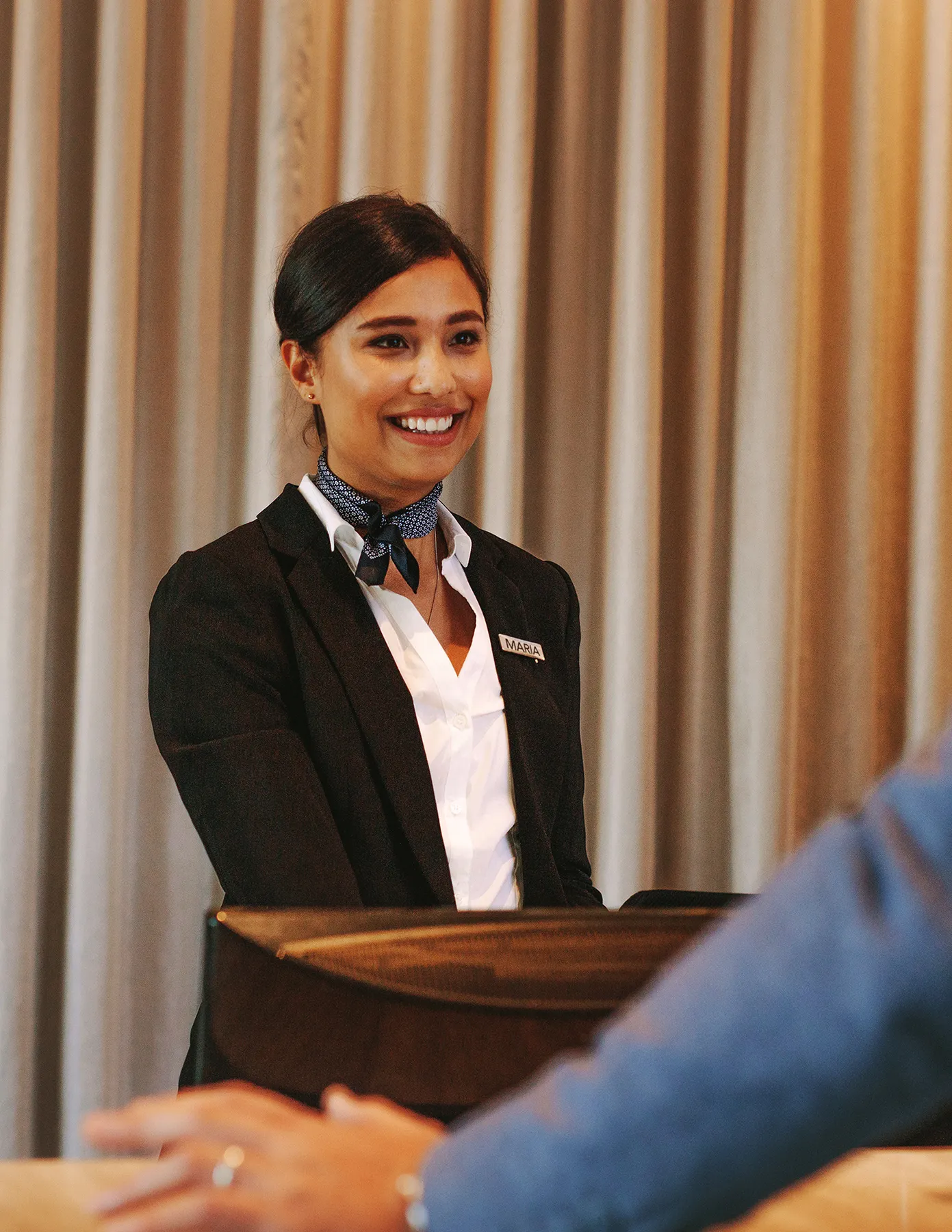 female-hotel-associate-maria-podium-greeting-guests-hmg-plus. Smiling event hostess standing at a check-in podium while greeting a guest at an indoor reception