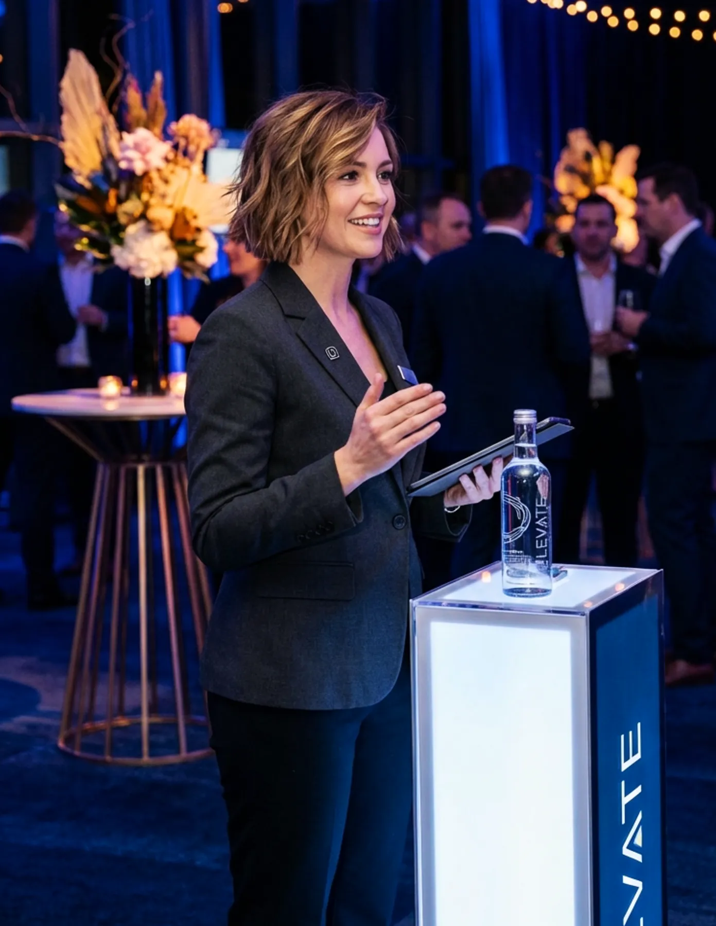 event-host-elevate-branded-podium-corporate-cocktail-event-hmg-plus. A female event host in a gray blazer speaking at an illuminated Elevate branded podium at a corporate cocktail event.