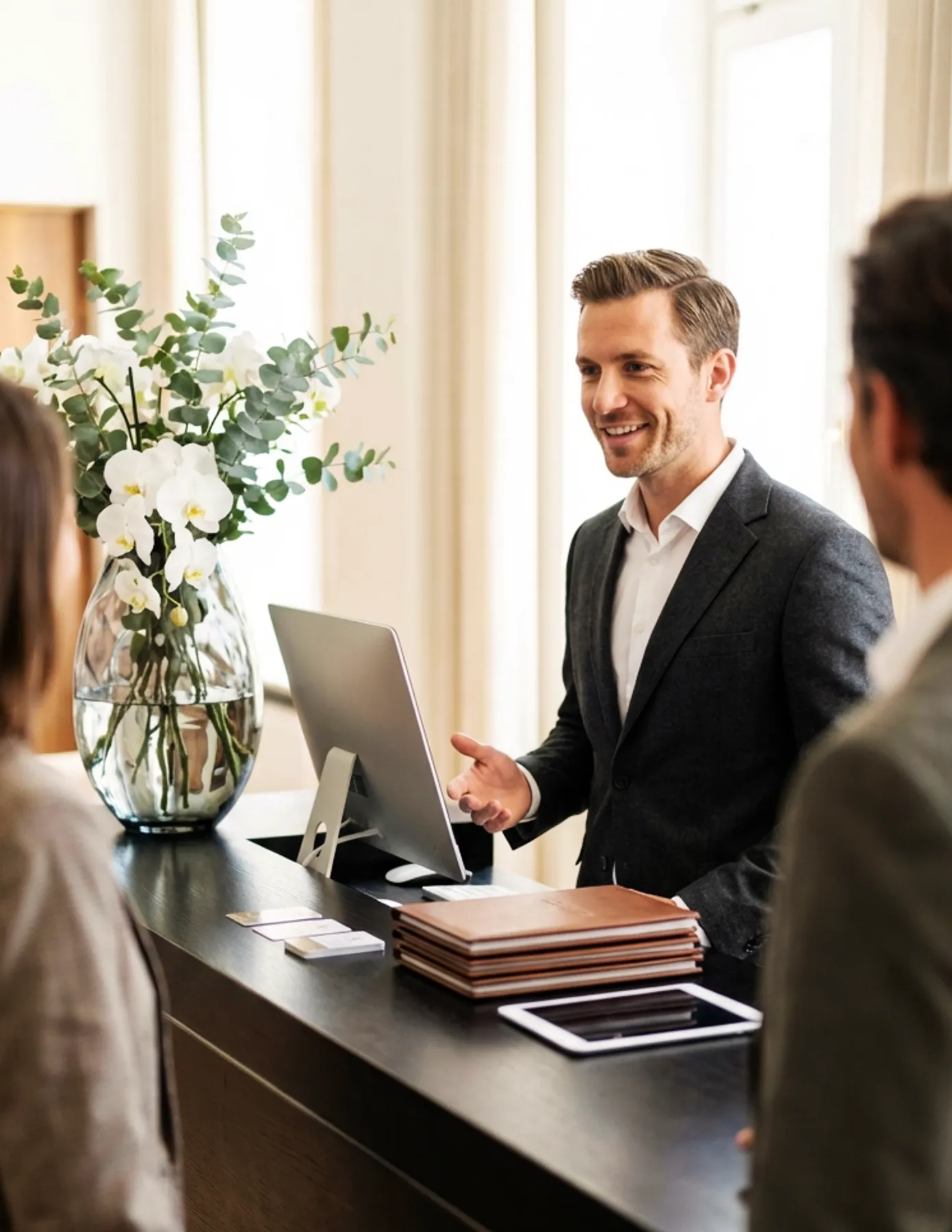 hotel-concierge-assisting-guests-front-desk-orchid-hmg-plus. A male hotel concierge smiling and assisting two guests at a sleek front desk with an orchid arrangement nearby.