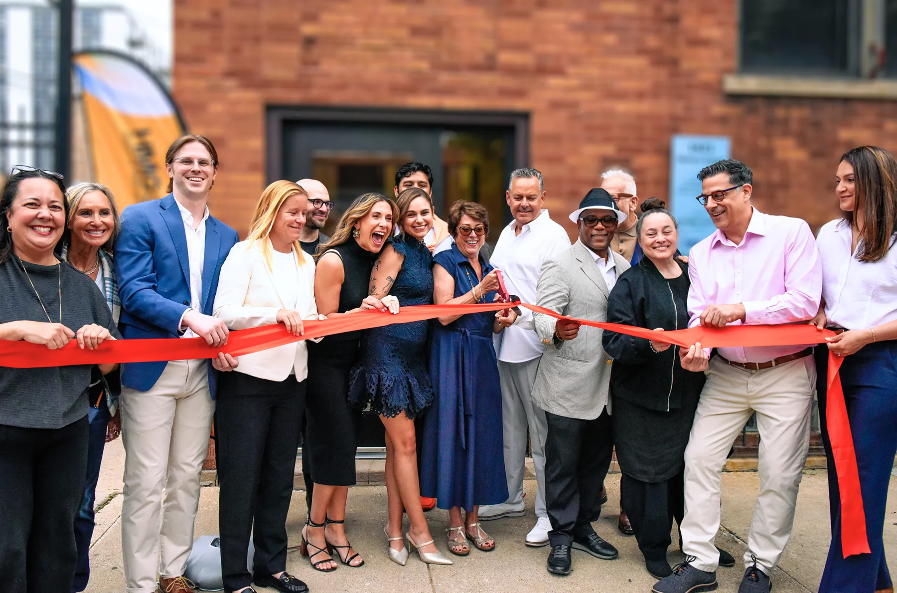 group-ribbon-cutting-grand-opening-ceremony-brick-building-hmg-plus. A large group of smiling people cutting a red ribbon together at a grand opening ceremony outside a brick building.