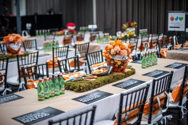 elegantly-decorated-event-banquet-tables-with-orange-florals-hmg-plus. An elegantly styled event banquet hall with long tables set with black Chiavari chairs, green glass water bottles, black printed place cards, orange floral centerpieces atop moss-covered risers, and orange and white striped gift bags tied to chair backs.