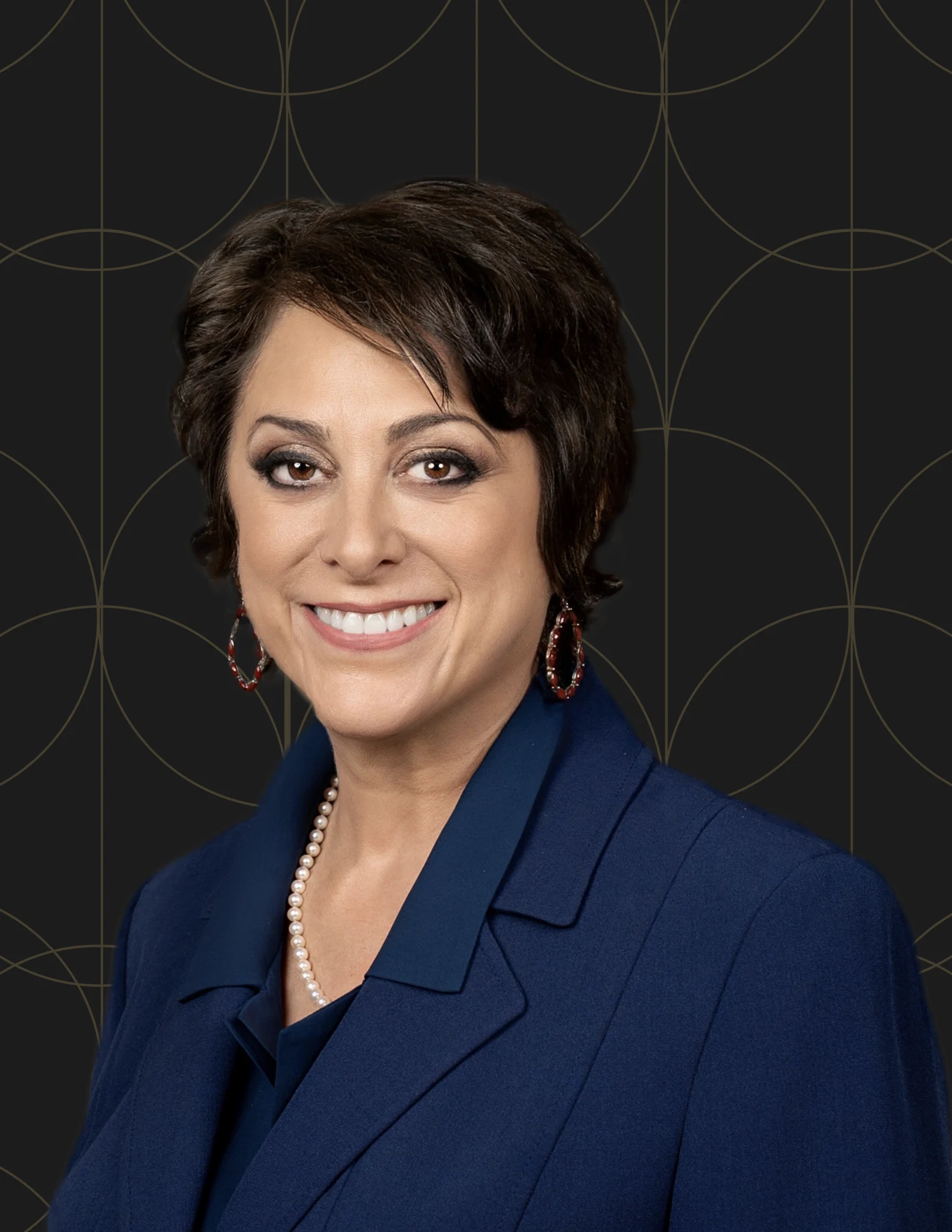 hmg-plus-team-member-headshot-woman-navy-blazer-pearls-hmg-plus. Professional headshot of a woman with short dark hair wearing a navy blazer, pearl necklace, and red hoop earrings.
