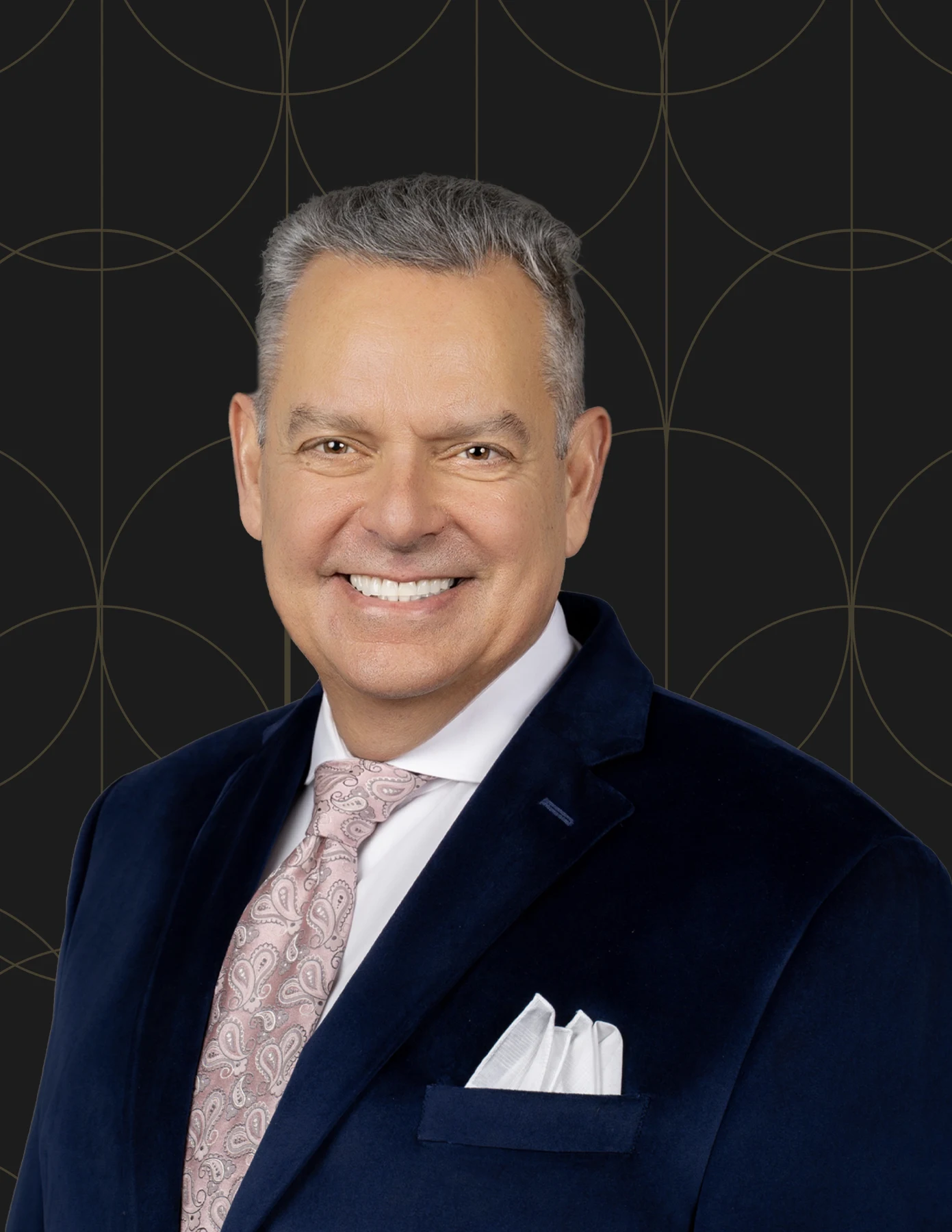 hmg-plus-team-member-headshot-man-navy-blazer-paisley-tie-hmg-plus. Professional headshot of a man with silver hair wearing a navy velvet blazer, pink paisley tie, and white pocket square.