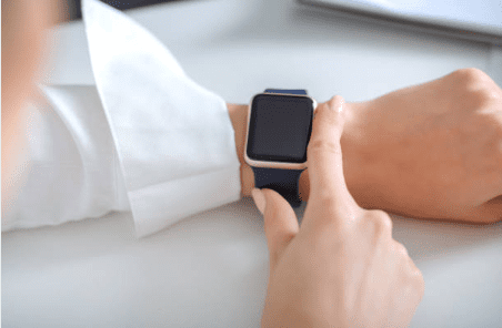 person-tapping-smartwatch-on-wrist-in-white-dress-shirt-hmg-plus. A person in a white shirt tapping the screen of a smartwatch with a navy blue band on their wrist, against a clean white desk surface.