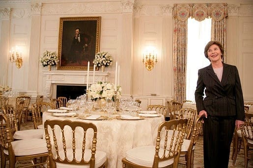 woman-smiling-in-elegantly-set-formal-dining-room-with-gold-chairs-hmg-plus. A woman in a dark pinstripe suit smiles while standing in an elegantly appointed formal dining room
