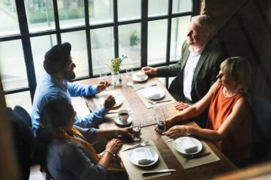 four-adults-engaged-in-conversation-hmg-plus. An overhead view of four adults engaged in conversation around a rustic wooden dining table set with plates, wine glasses, and coffee cups, beside large windows in a warmly lit restaurant.